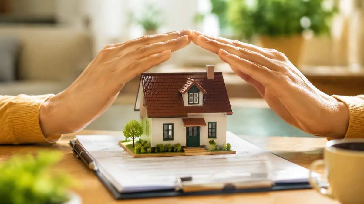Casa em miniatura protegida por mãos sobre documentos de apólice na mesa