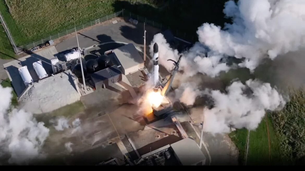 overhead view of a black and white rocket launching from a pad surrounded by grassy fields 